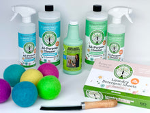 Load image into Gallery viewer, closer view of back row hows two large bottles of all-purpose cleaning solution with two spray bottles next to them, in the middle is a bottle of green hard water stain remover, on the left front is 6 multi-colored dryer balls, front right is a flat rectangular box of laundry detergent sheets and in the very front is a flat black brush with brown wooden handle