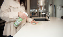 Load image into Gallery viewer, women next to a kitchen counter with sink, spraying pur evergreen all-purpose cleaning and wiping counter with Swedish dishcloth