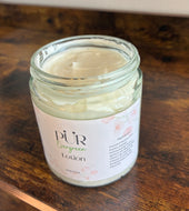 Jar of lotion labeled 'PUR Evergreen Lotion' on a wooden surface. This glass jar is 140 oz. This lotion is made with coconut butter, coco butter, mango butter, jojoba butter, apricot oil, rosehip oil, vitamin e, glycerin, and vitamin c. It is hydrating without being greasy or leaving behind any residue.