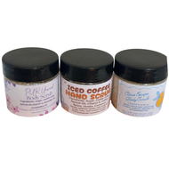 Three small jars of body and hand scrubs lined up against a white background. The left jar is labeled 'PURified Body Scrubs', the middle jar is labeled 'ICED COFFEE HAND SCRUB', and the right jar is labeled 'Citrus Zing Body Scrub'.